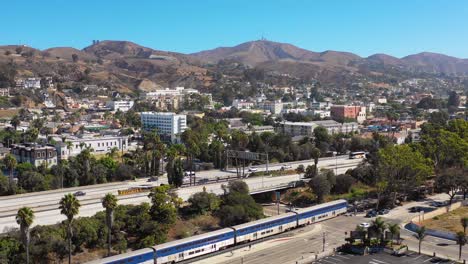 A-drone-aerial-of-the-Pacific-Surfline-Amtrak-train-passing-through-the-Southern-California-beach-town-of-Ventura-California-with-freeway-foreground-and-mountains-background
