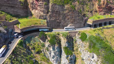 An-aerial-shot-of-a-convoy-of-busses-traveling-on-a-dangerous-narrow-mountain-road-along-the-ocean-Chapmans-Peak-Road-near-Cape-Town-South-Africa-2