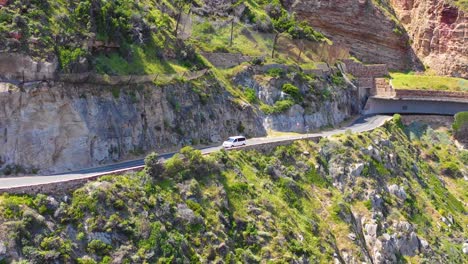 An-aerial-shot-of-a-car-minivan-traveling-on-a-dangerous-narrow-mountain-road-along-the-ocean-Chapmans-Peak-Road-near-Cape-Town-South-Africa