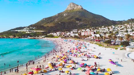 Spectacular-aerial-over-a-crowded-and-busy-holiday-beach-at-Camps-Bay-Cape-Town-South-Africa-with-Lion's-Head-mountain-background
