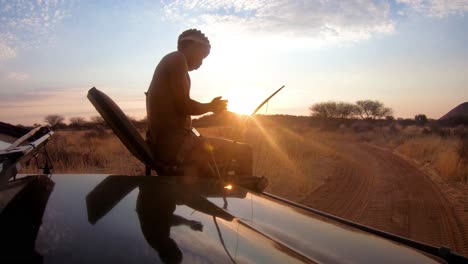 A-native-spotter-guide-sits-on-the-front-of-a-safari-jeep-vehicle-spotting-wildlife-on-the-plains-of-Africa-at-sunset
