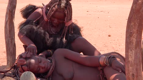 Eine-Himba-Frau-Hat-Ihre-Erstaunlichen-Geflochtenen-Und-Schlammverkrusteten-Dreadlocks-Haare-Von-Einer-Anderen-Frau-In-Einem-Kleinen-Dorf-An-Der-Grenze-Zu-Namibia-Und-Angola-Gestylt-1