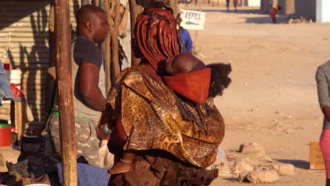Eine-Himba-Stammesfrau-Mit-Baby-Auf-Dem-Rücken-Und-Erstaunlicher-Frisur-Aus-Schlamm-Und-Zöpfen-Und-Dreadlocks-In-Der-Marktstadt-Opuwo,-Namibia