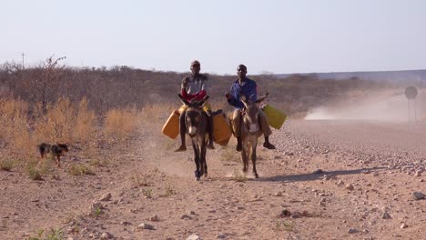 Two-Himba-men-ride-donkeys-along-a-dusty-road-in-Africa-Damaraland-Namibia-bringing-water-to-remote-villages