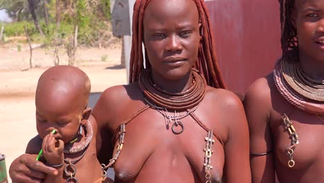 Schöne-Himba-Afrikanische-Stammesfrauen-Halten-Ihre-Babys-und-Posieren-Mit-Schlammfrisur-und-Dreadlocks-und-Runden-Halsketten