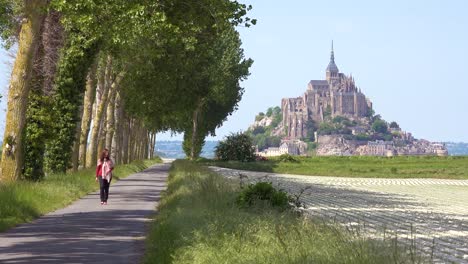 A-woman-walks-on-a-pretty-road-through-the-countryside-with-Mont-Saint-Michel-monastery-island-in-distance-Normandy-France