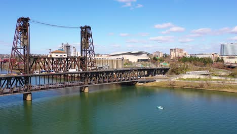 Bonita-Antena-Del-Tren-De-Pasajeros-De-Amtrak-Cruzando-El-Puente-Y-Llegando-A-Portland,-Oregón,-Con-El-Tren-Ligero-Arriba-Y-El-Centro-De-Moda-De-Fondo