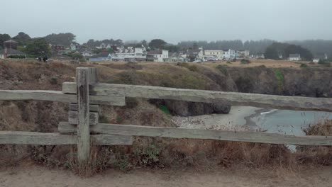 Pan-shot-of-sturdy-wooden-fence-people-a-small-beach-and-cove-and-the-historic-wooden-buildings-of-Mendicino-CA