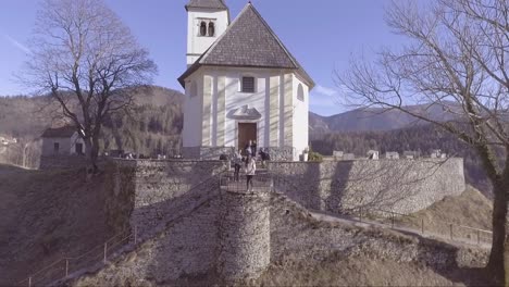 Aerial-of-a-small-white-church-in-the-mountains-of-Slovenia-and-woman-and-photographers