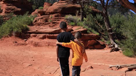 Un-Niño-Indio-Americano-Nativo-Y-Su-Hermano-Caminan-Del-Brazo-Cerca-De-Algunas-Colinas-De-Red-Rock-En-Sedona,-Arizona