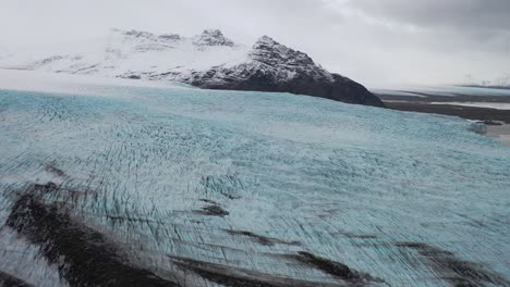 Antenne-Eines-Gletschers,-Der-Von-Einer-Schneebedeckten-Bergkette-In-Island-Eingefroren-Ist