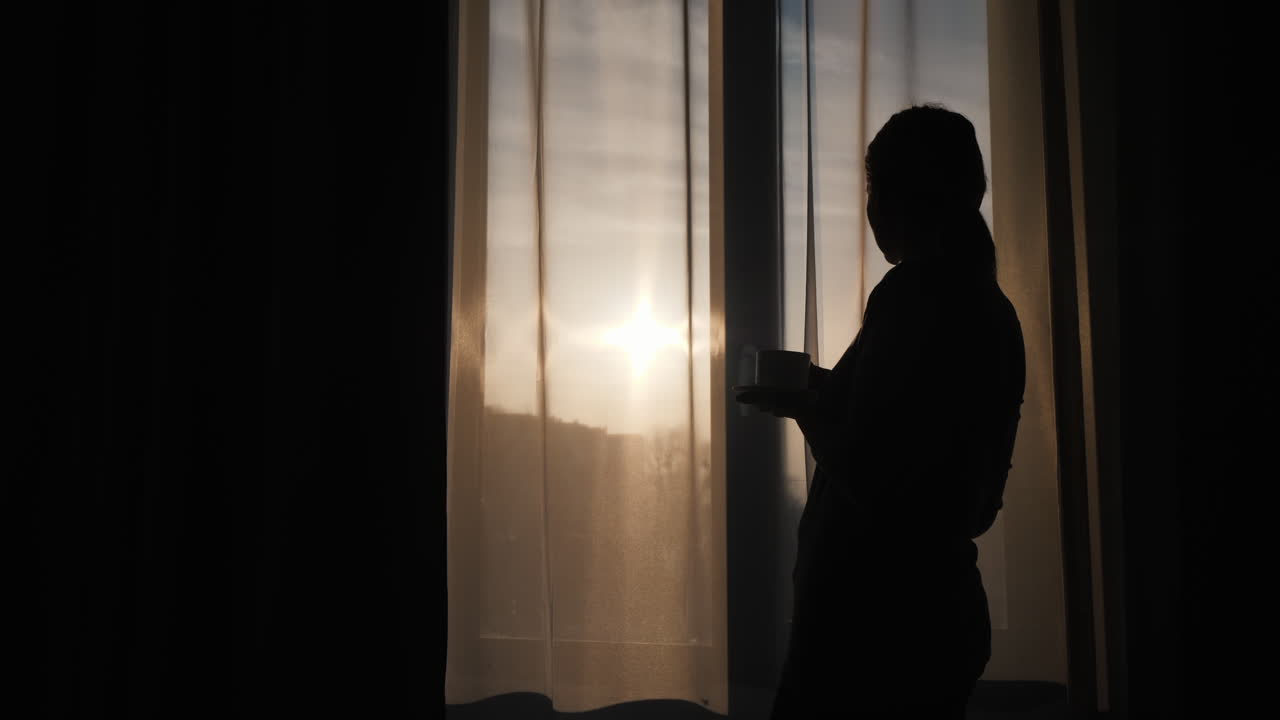 Premium stock video - The silhouette of a woman at the window drinks a ...