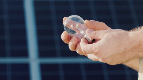 Side-View-Of-A-Man's-Hands-Holding-A-Light-Bulb-Against-The-Background-Of-Solar-Panels