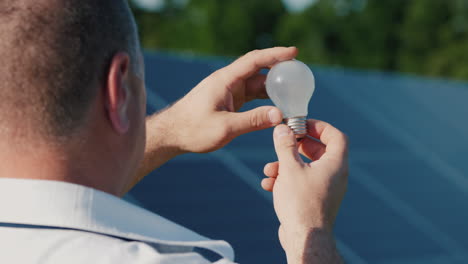 A-Man-Looks-At-An-Electric-Light-Bulb-Against-The-Background-Of-Solar-Power-Plant-Panels