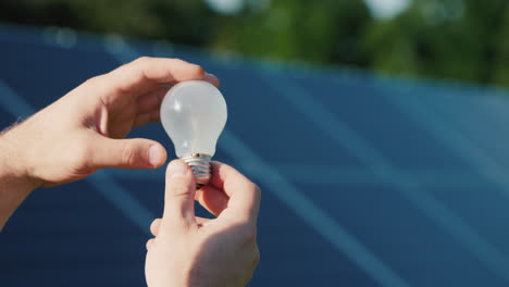 Man's-Hands-Hold-A-Light-Bulb-Against-The-Background-Of-Solar-Panels