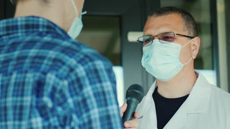 A-Reporter-In-A-Protective-Mask-Interviews-A-Doctor-At-The-Entrance-To-The-Hospital