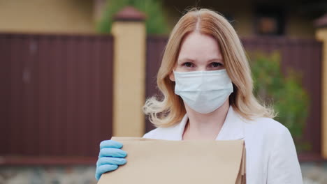 A-Portrait-Of-A-Masked-Woman-Holding-Bags-Of-Groceries-Stands-Near-Her-House