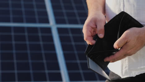 A-Man-Holds-An-Empty-Purse-Against-The-Background-Of-Solar-Panels