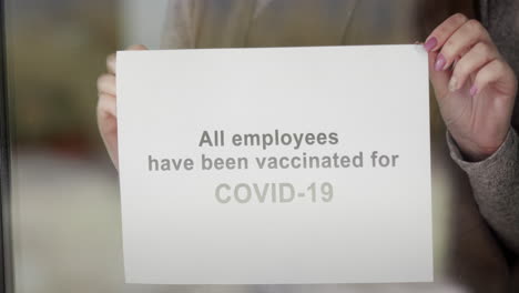 An-Employee-Hangs-An-Announcement-On-The-Door-Of-The-Establishment-That-All-Employees-Are-Vaccinated-And-Visiting-Safely