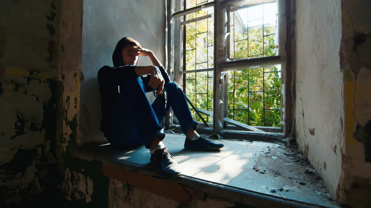 Premium stock video - Teenager with a gun sits in an abandoned building 2