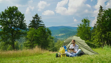 Ein-Junger-Männlicher-Tourist-Benutzt-Einen-Laptop-Beim-Camping-In-Der-Nähe-Eines-Zeltes-1